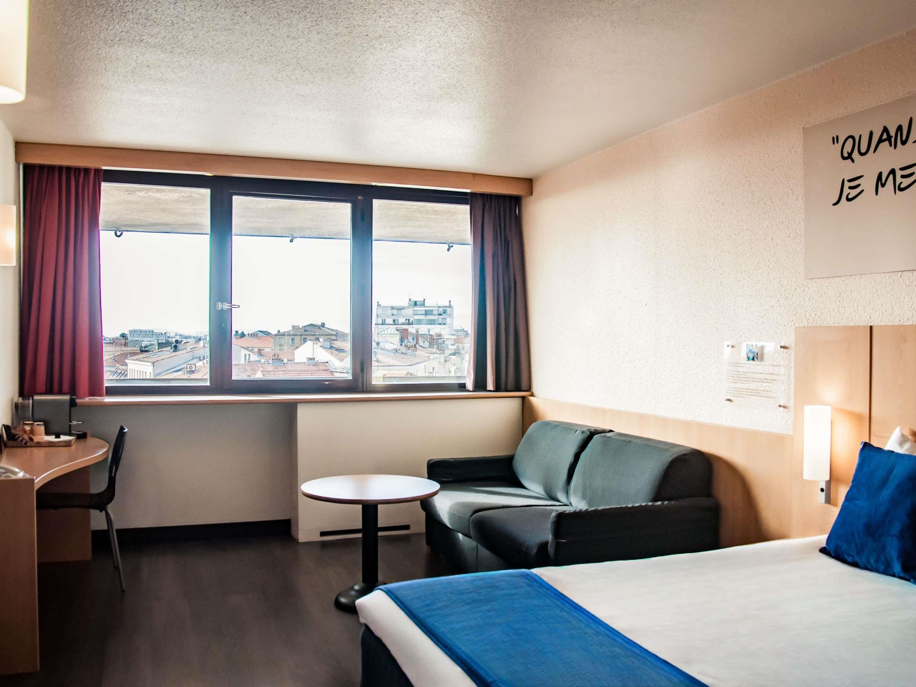 Habitación de hotel con cama, escritorio, silla, una mesita y ventana con cortinas.