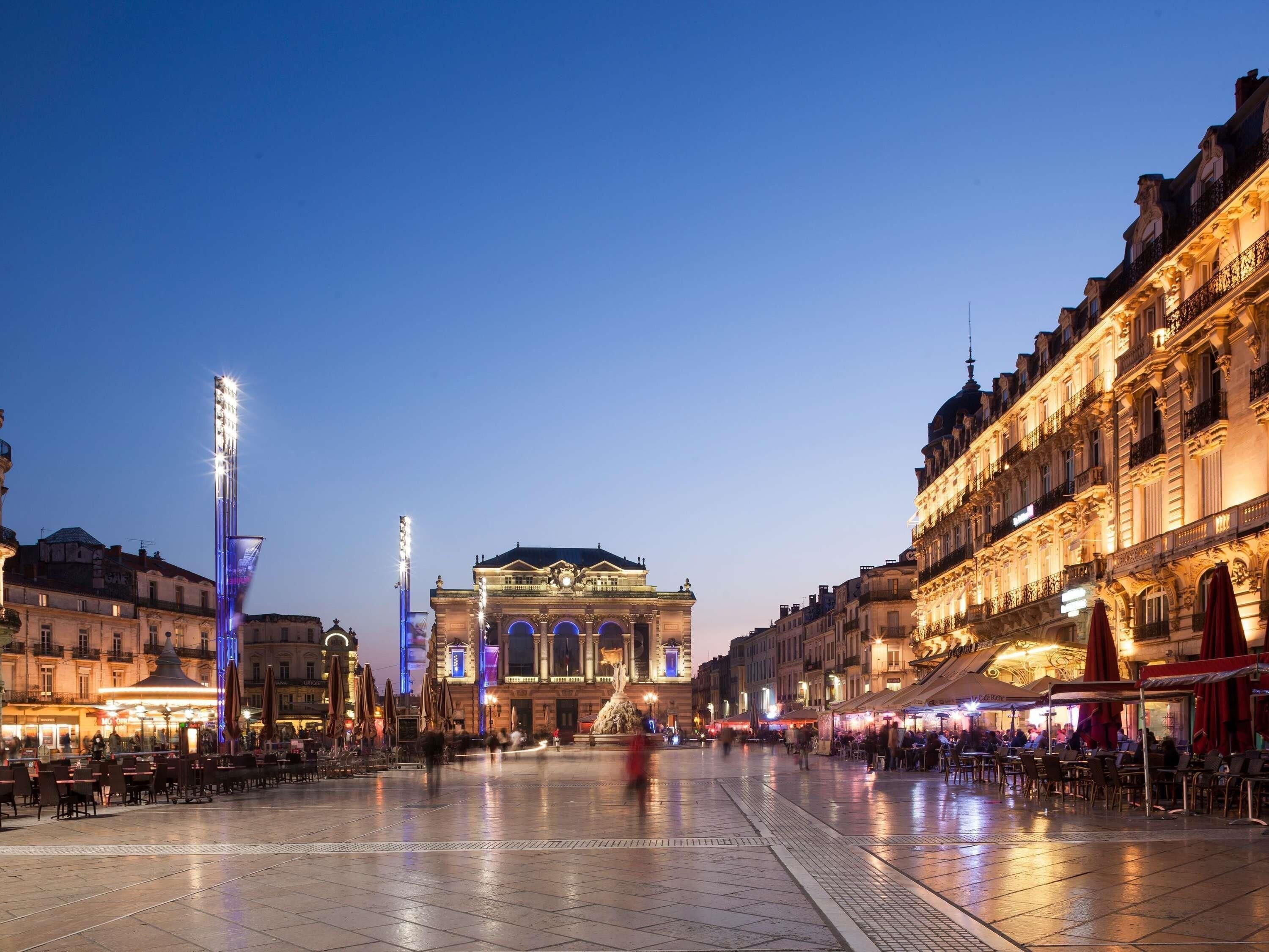 A spacious square with a prominent building featuring classical architecture, surrounded by cafes and outdoor seating.