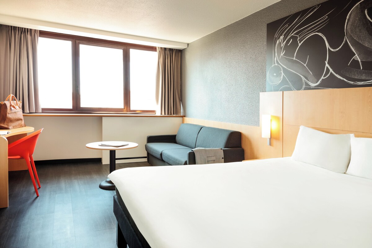 A hotel room with a bed, a sofa, a small table, a chair, and a window with curtains.