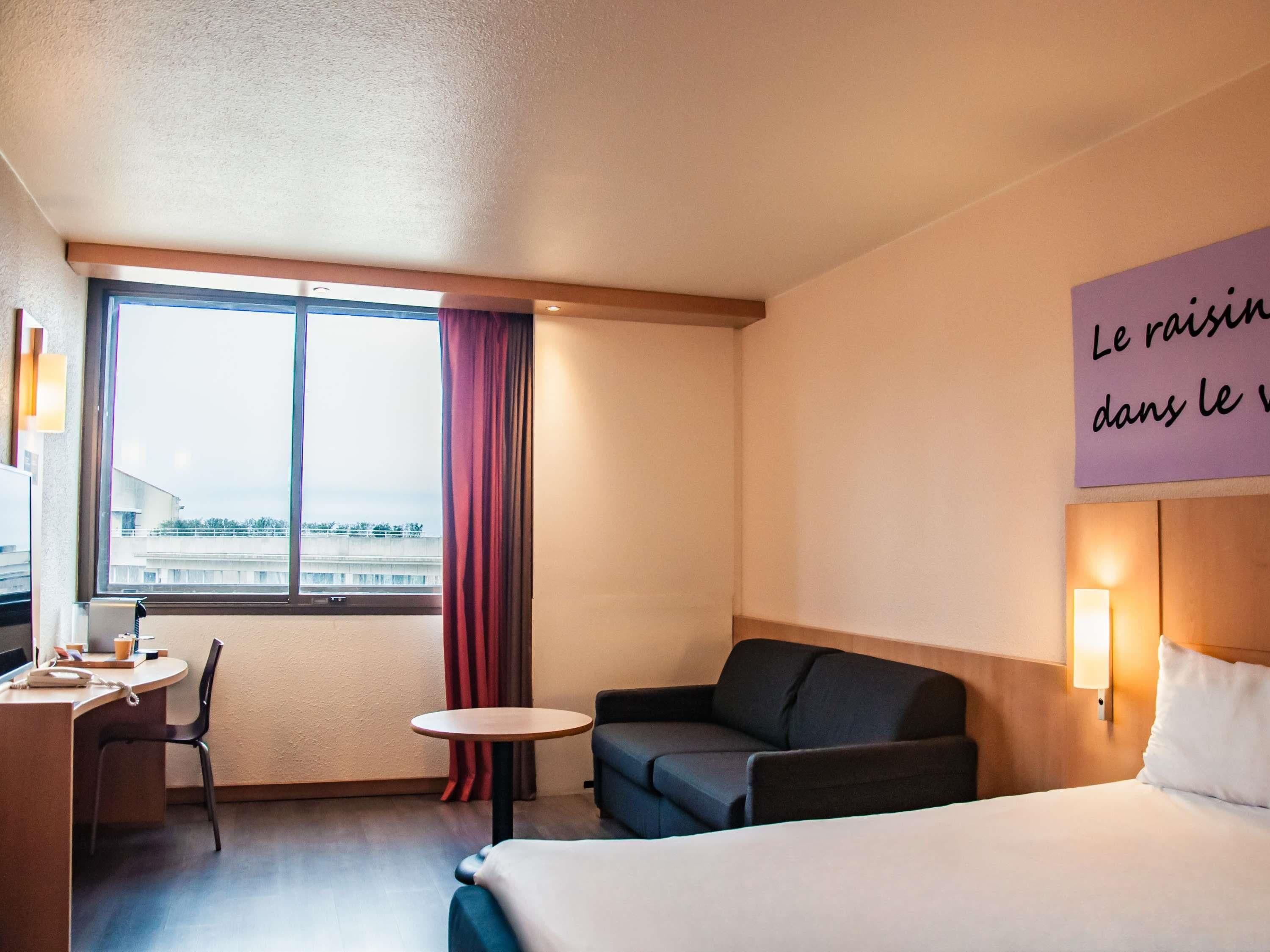 Habitación de hotel con cama, escritorio, silla, mesita y sofá. Se ve una ventana con vistas a edificios y una cortina.