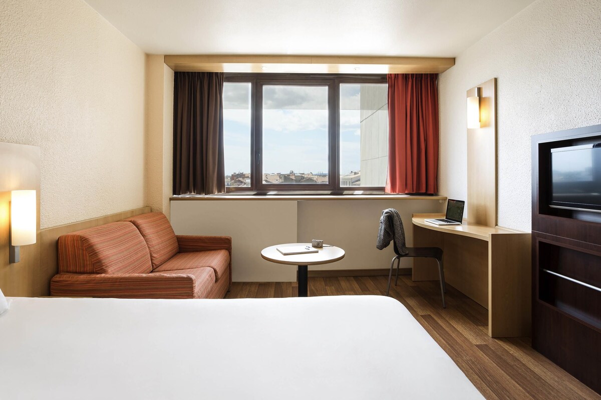 Una habitación de hotel con una cama, un sofá, un escritorio con una computadora portátil, una silla, una mesita y un televisor.
