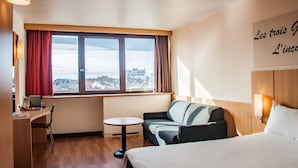 A hotel room with a bed, a sofa, a small table, a desk, and a window with curtains.