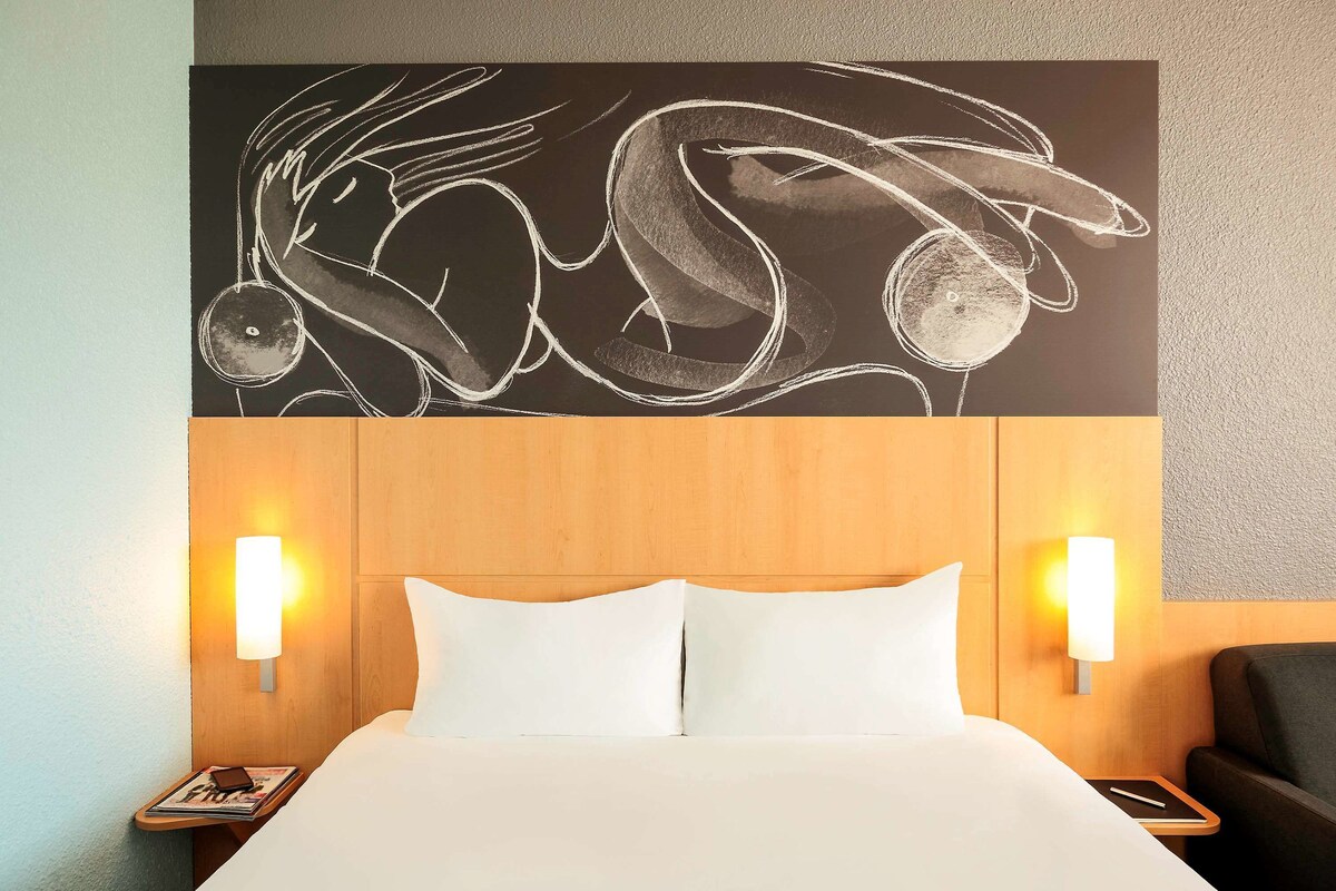 A hotel room with a large bed, two bedside lamps, a wooden headboard, and a wall art piece above the bed.