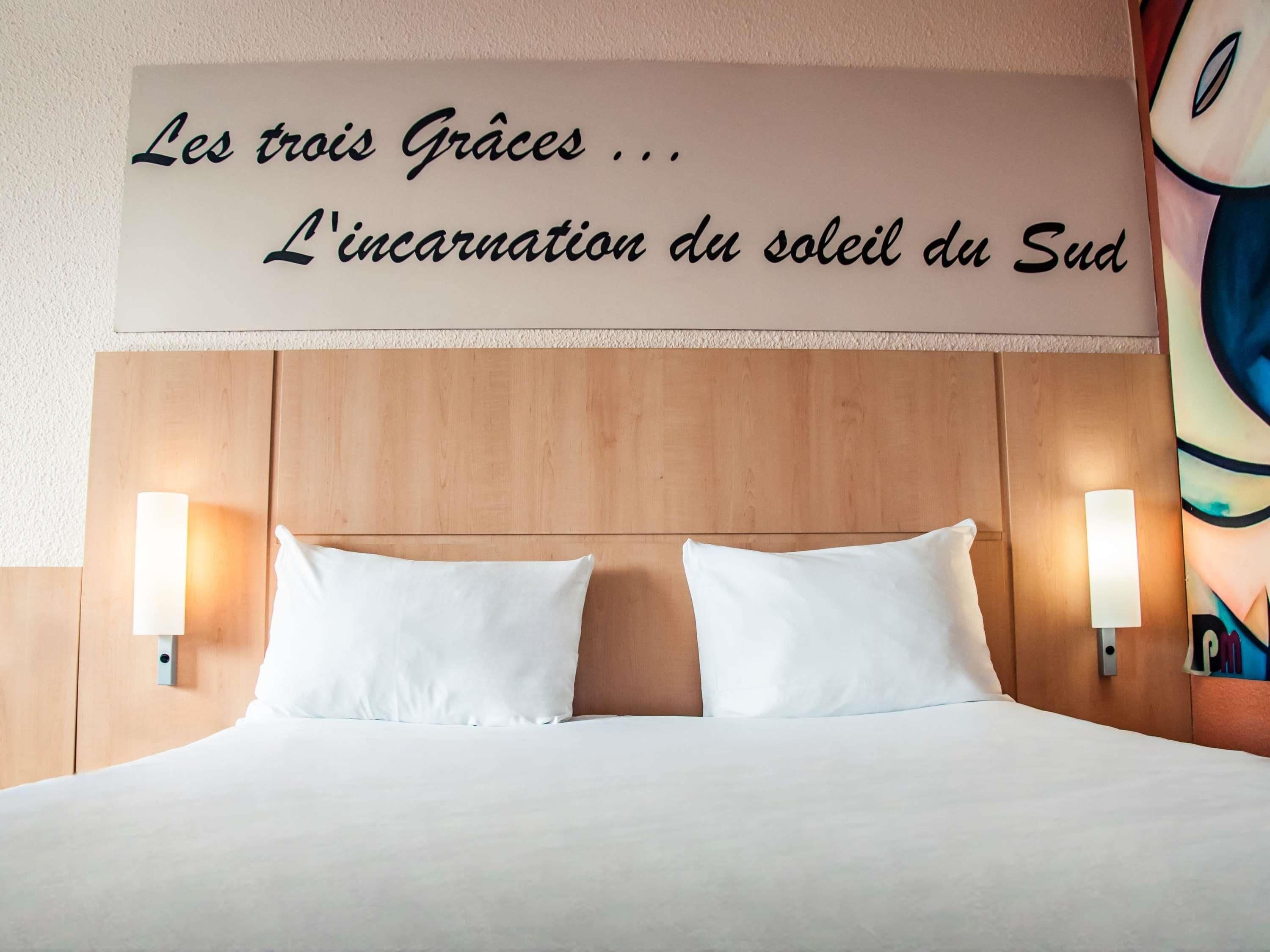 Habitació d'hotel amb un capçal de llit de fusta, dos coixins blancs i làmpades fixades a la paret. Un cartell sobre el llit diu "Les tres Gràcies... L'encarnació del sol del Sud.".