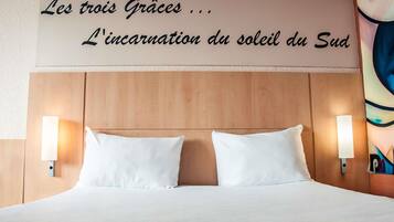 Habitació d'hotel amb un capçal de llit de fusta, dos coixins blancs i làmpades fixades a la paret. Un cartell sobre el llit diu "Les tres Gràcies... L'encarnació del sol del Sud.".
