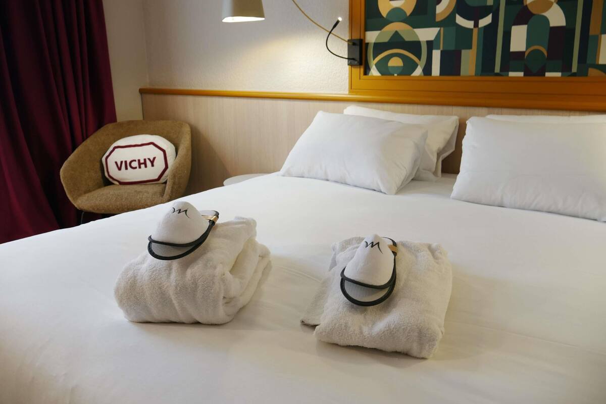 Una habitación de hotel con una cama bien hecha, una silla de la marca VICHY y un cuadro en la pared.