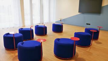 Une salle de conférence avec des chaises bleues et des tables rouges, un grand écran mural et une fenêtre avec des rideaux.