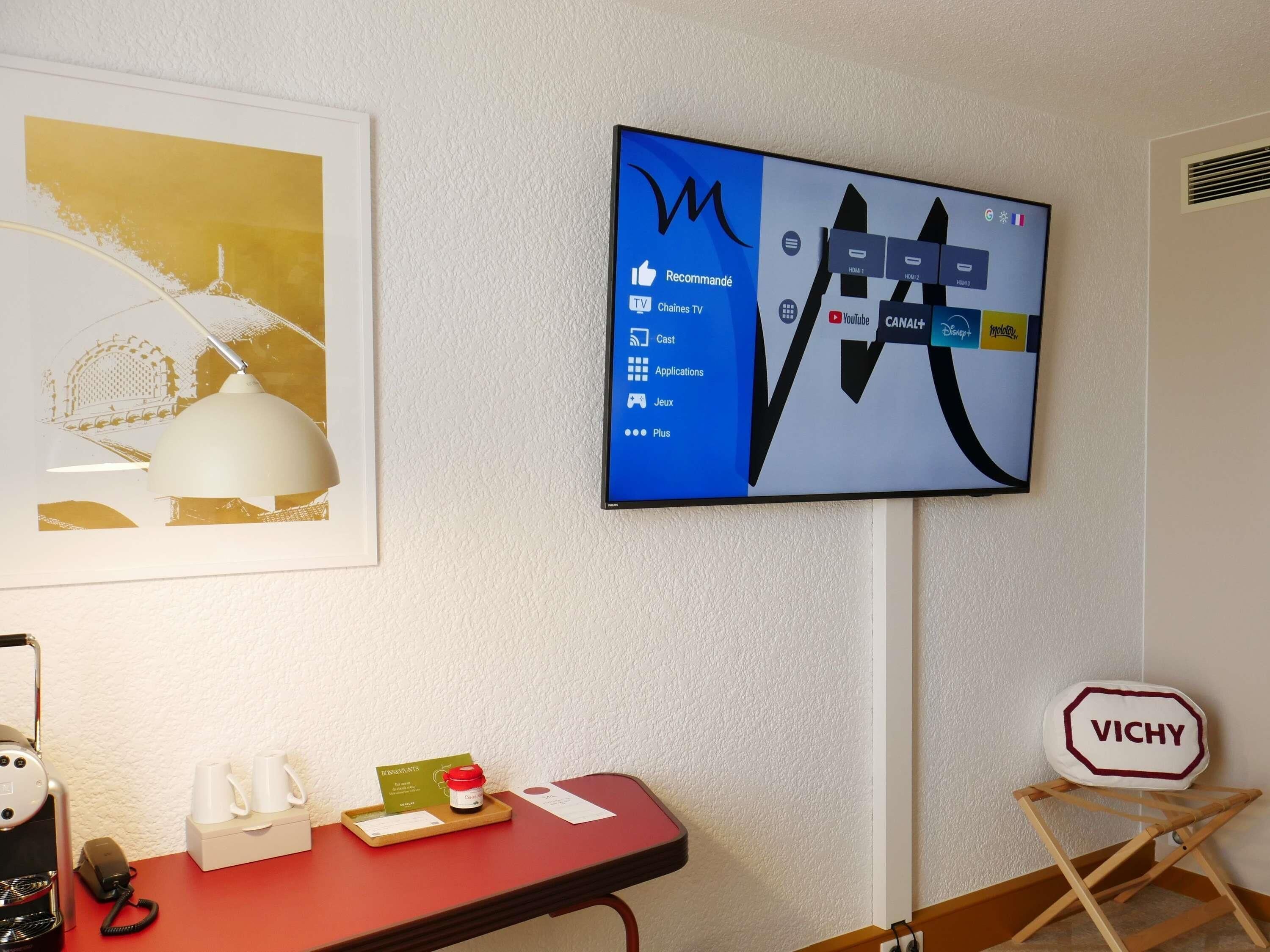 A hotel room with a mounted TV displaying a TV channel interface, a framed artwork on the wall, a desk with a coffee maker, a tray with a card and a small bottle, and a Vichy sign.
