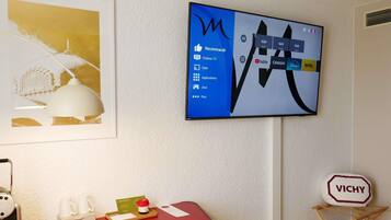 A hotel room with a mounted TV displaying a TV channel interface, a framed artwork on the wall, a desk with a coffee maker, a tray with a card and a small bottle, and a Vichy sign.