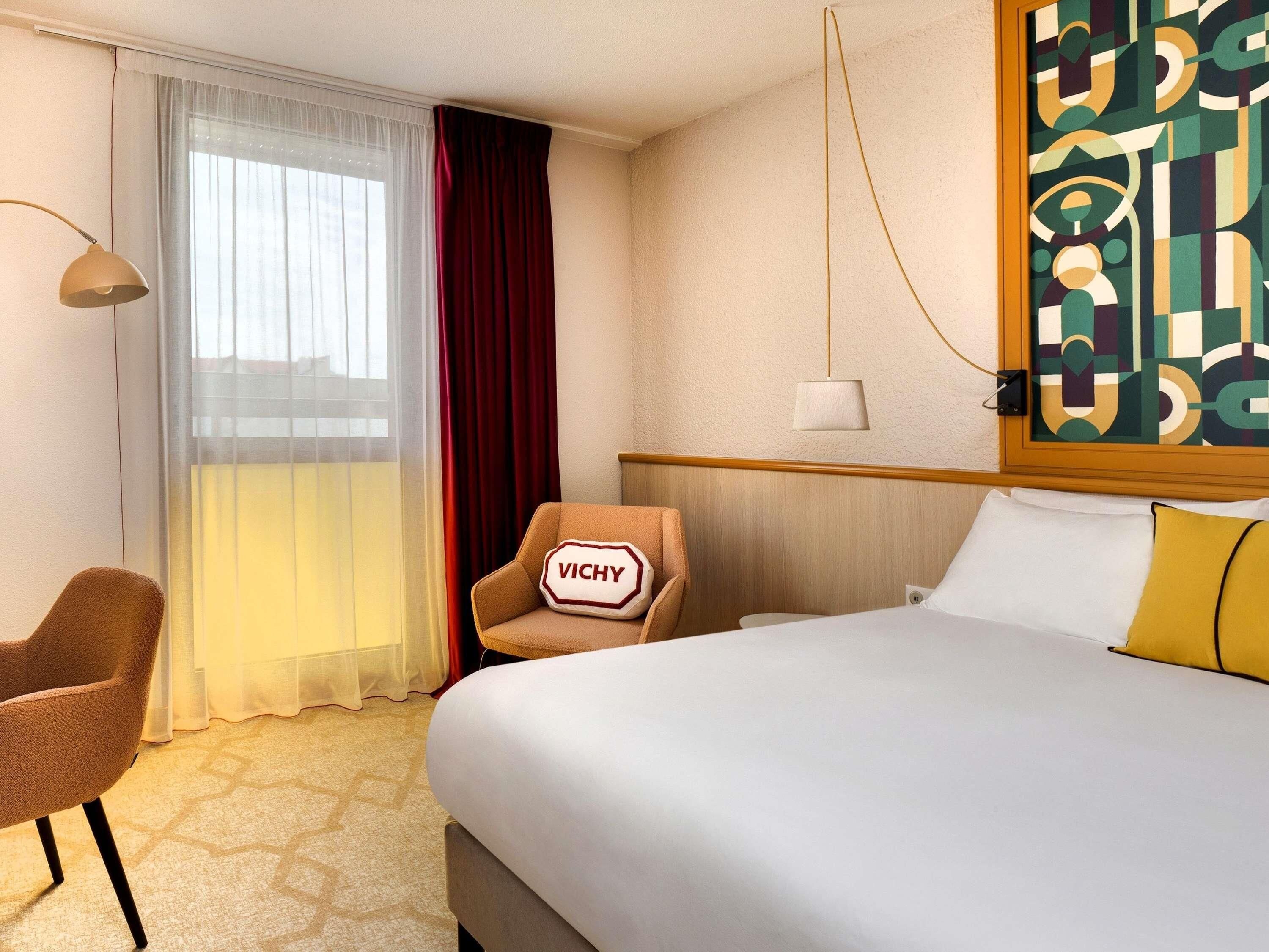 A hotel room with a bed, a chair, a desk, and a window with curtains.