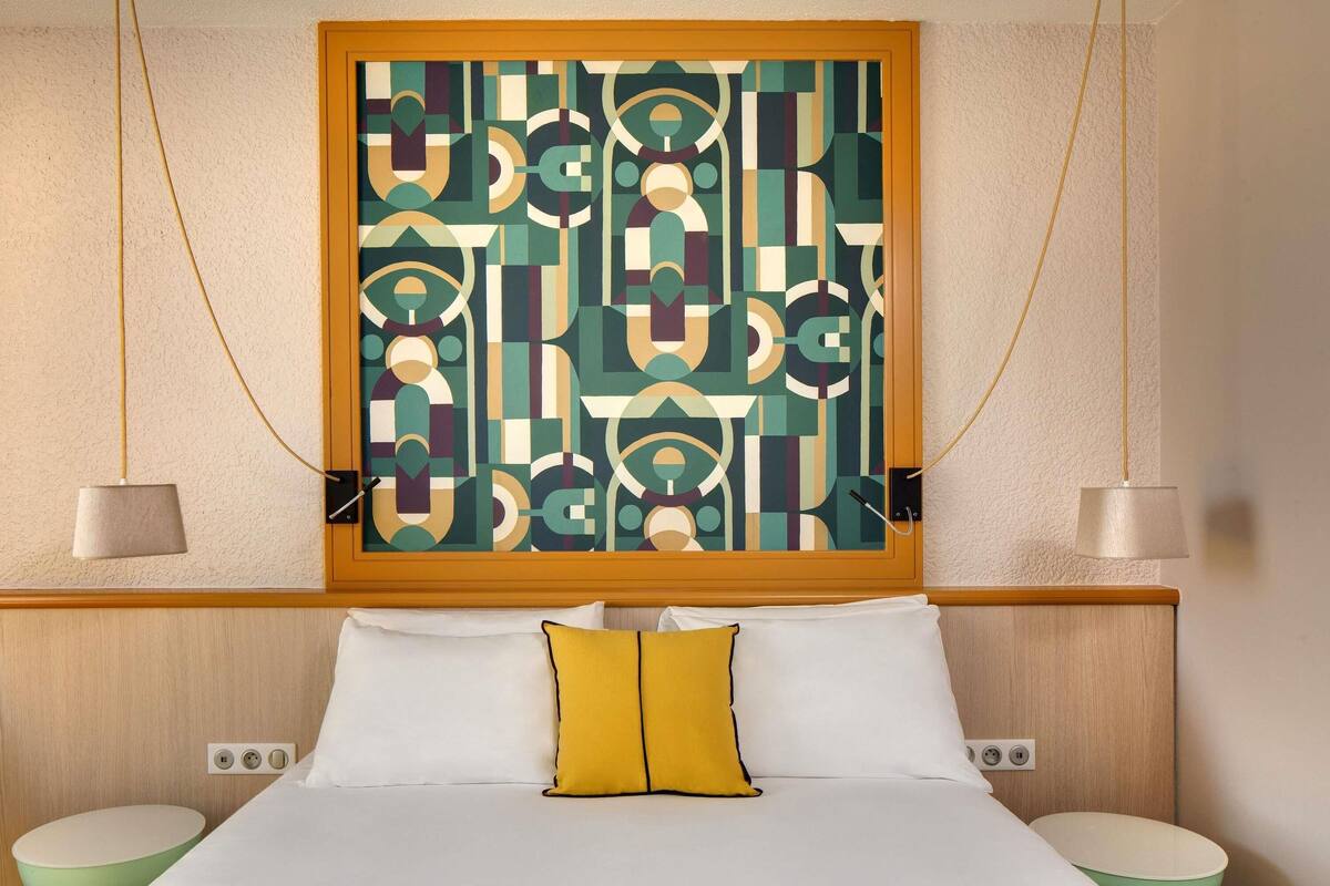 A hotel room with a bed, two bedside tables, a wall-mounted abstract painting, and a hanging lamp.