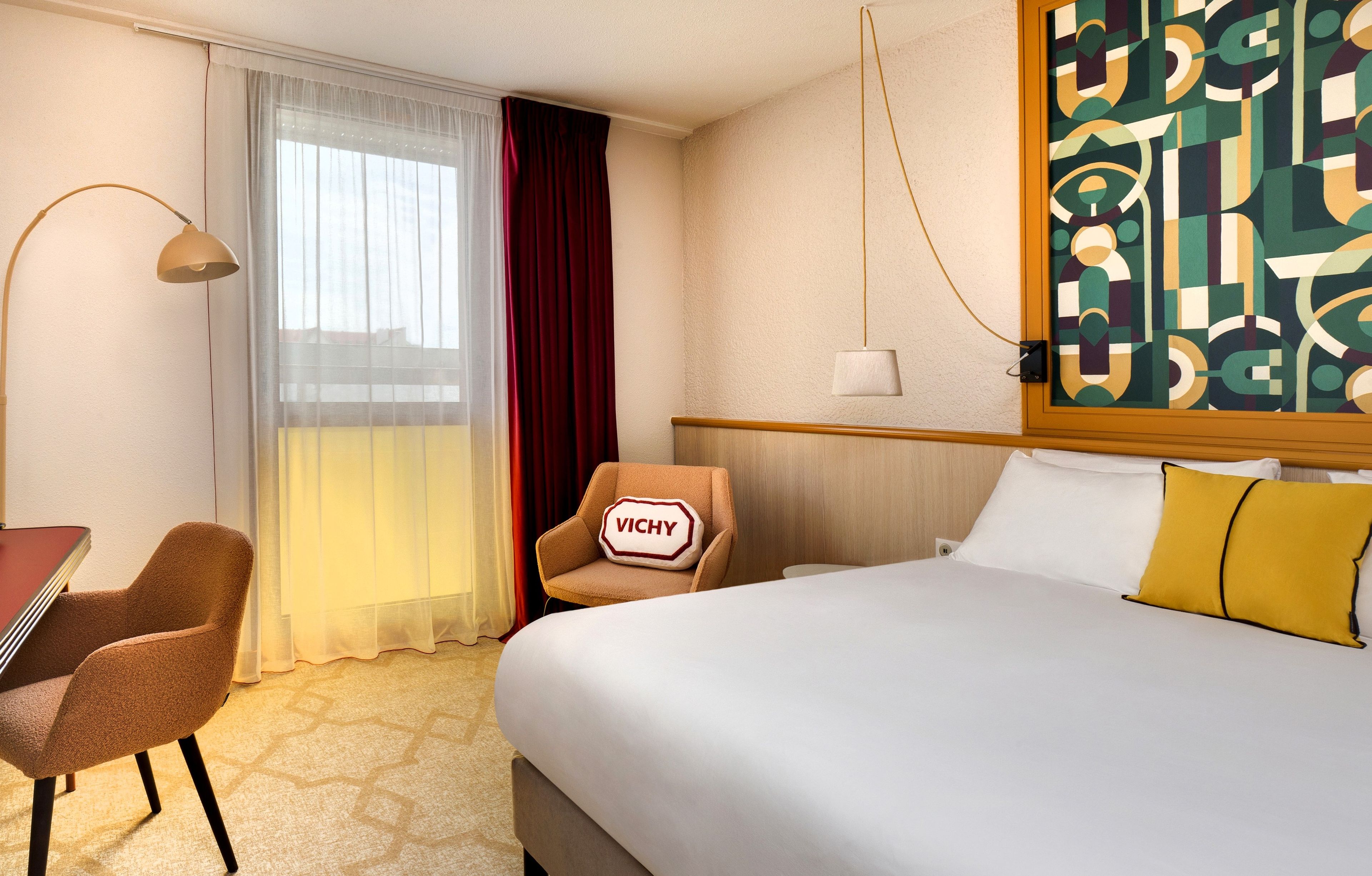 A hotel room with a bed, a desk, a chair, a window with curtains, and a painting on the wall.
