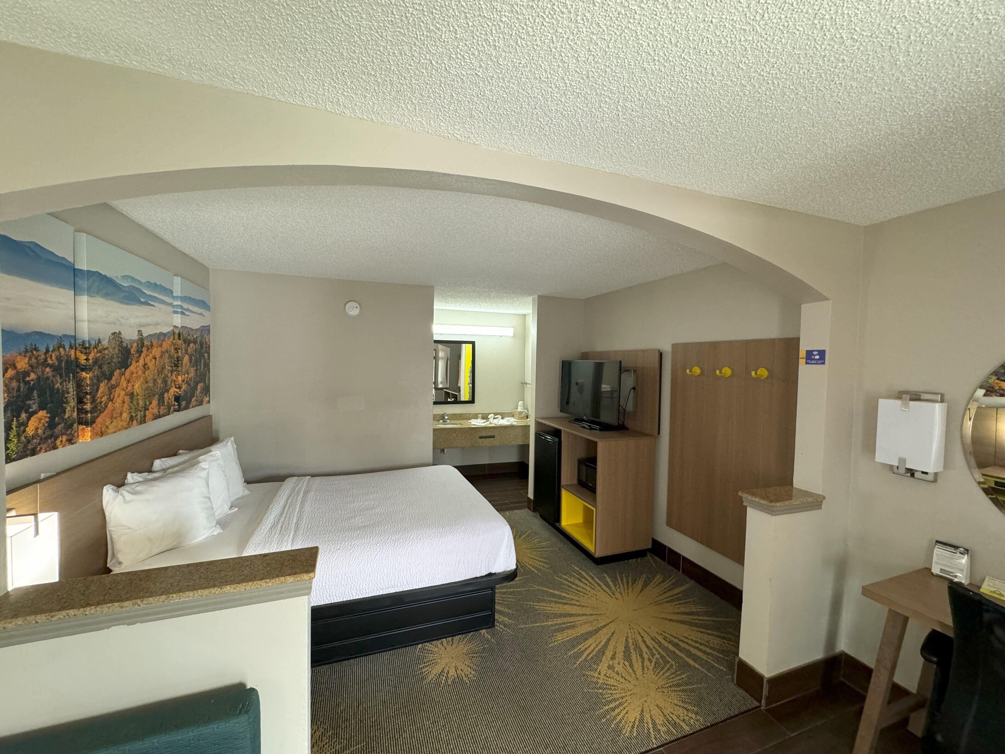A hotel room with a bed, a desk, a television, and a bathroom visible through an arched doorway.