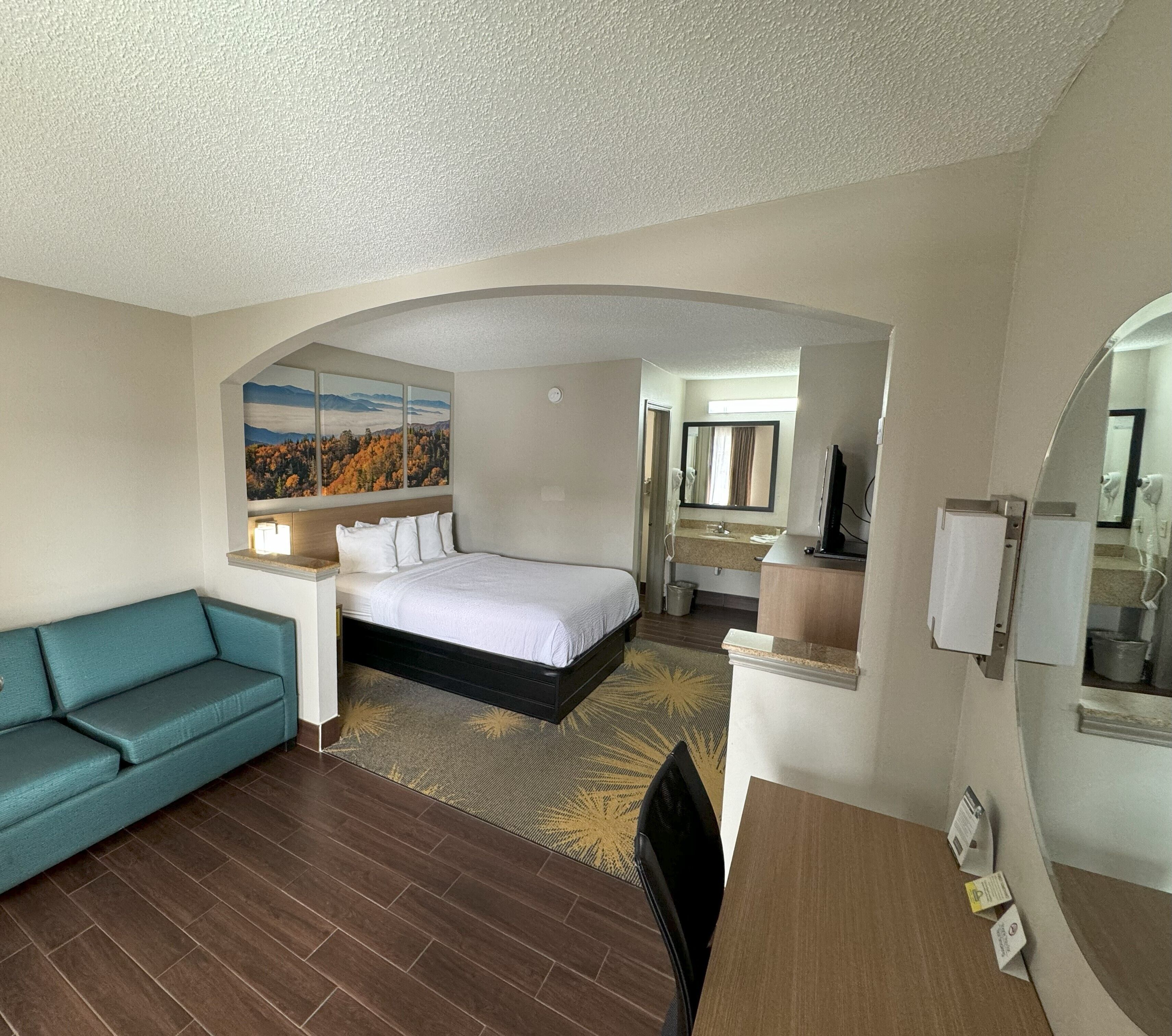 A hotel room with a bed, a sofa, a desk, a mirror, and a painting of a landscape.
