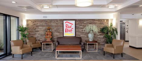 A lobby with a sofa, chairs, a coffee table, a wall-mounted Buddha statue, a 'Red Roof Plus' sign, and a stone wall.