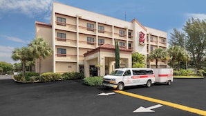 A multi-story hotel with a red roof sign, a parking lot with a white van, and palm trees. - Red Roof Inn PLUS+ Miami Airport (Miami)