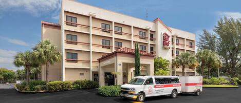 Un hotel de varios pisos con techo rojo, estacionamiento con una camioneta blanca y palmeras.