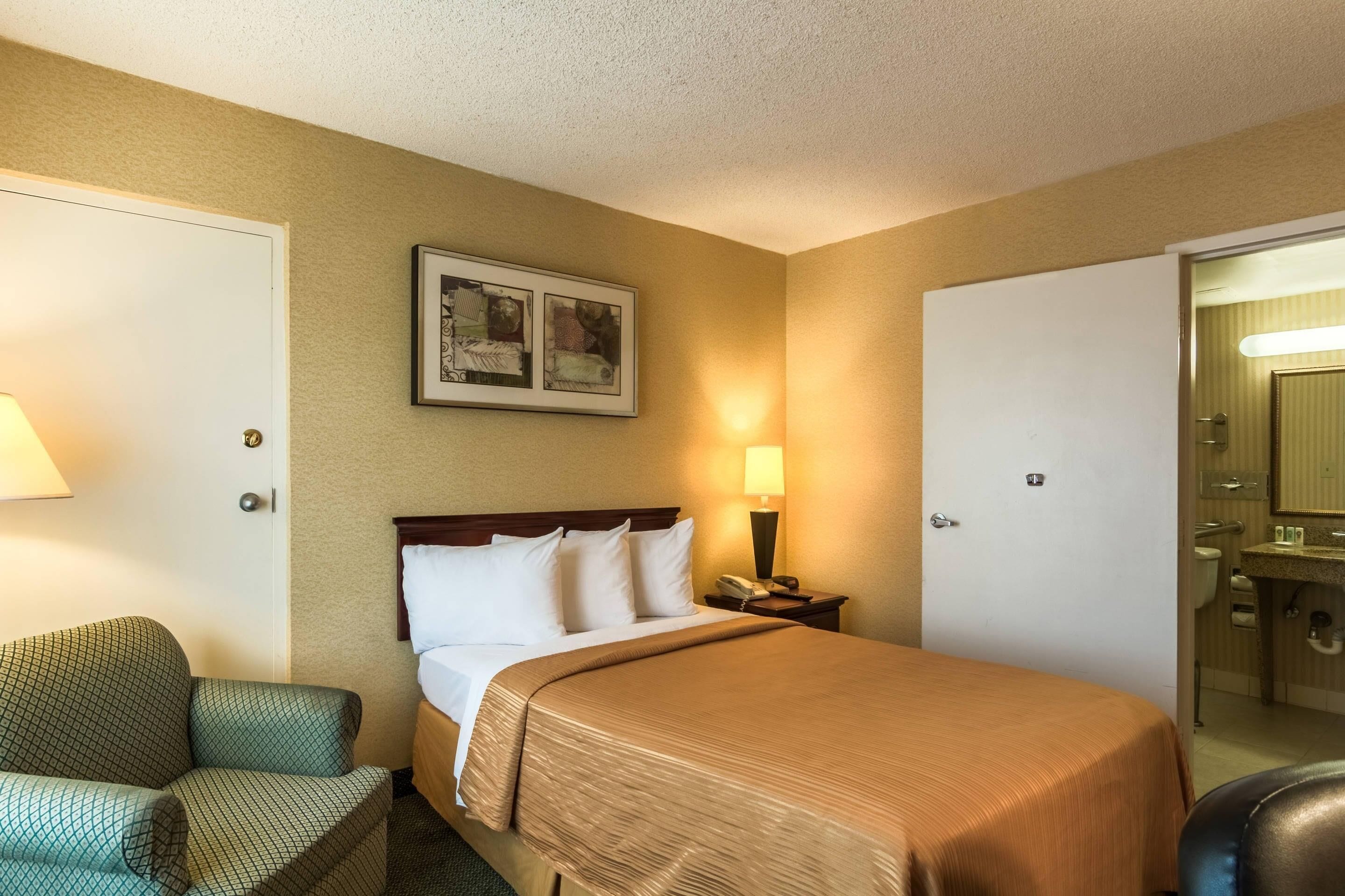 A hotel room with a bed, a chair, a bedside table with a lamp, a framed picture on the wall, and a door leading to a bathroom.