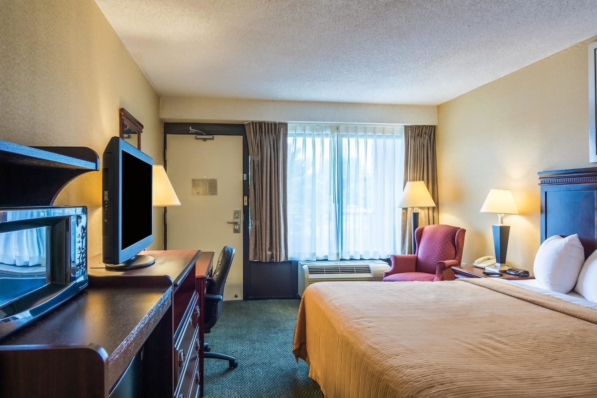 Quarto de hotel com cama, escrivaninha com televisão, cadeira e duas luminárias.
