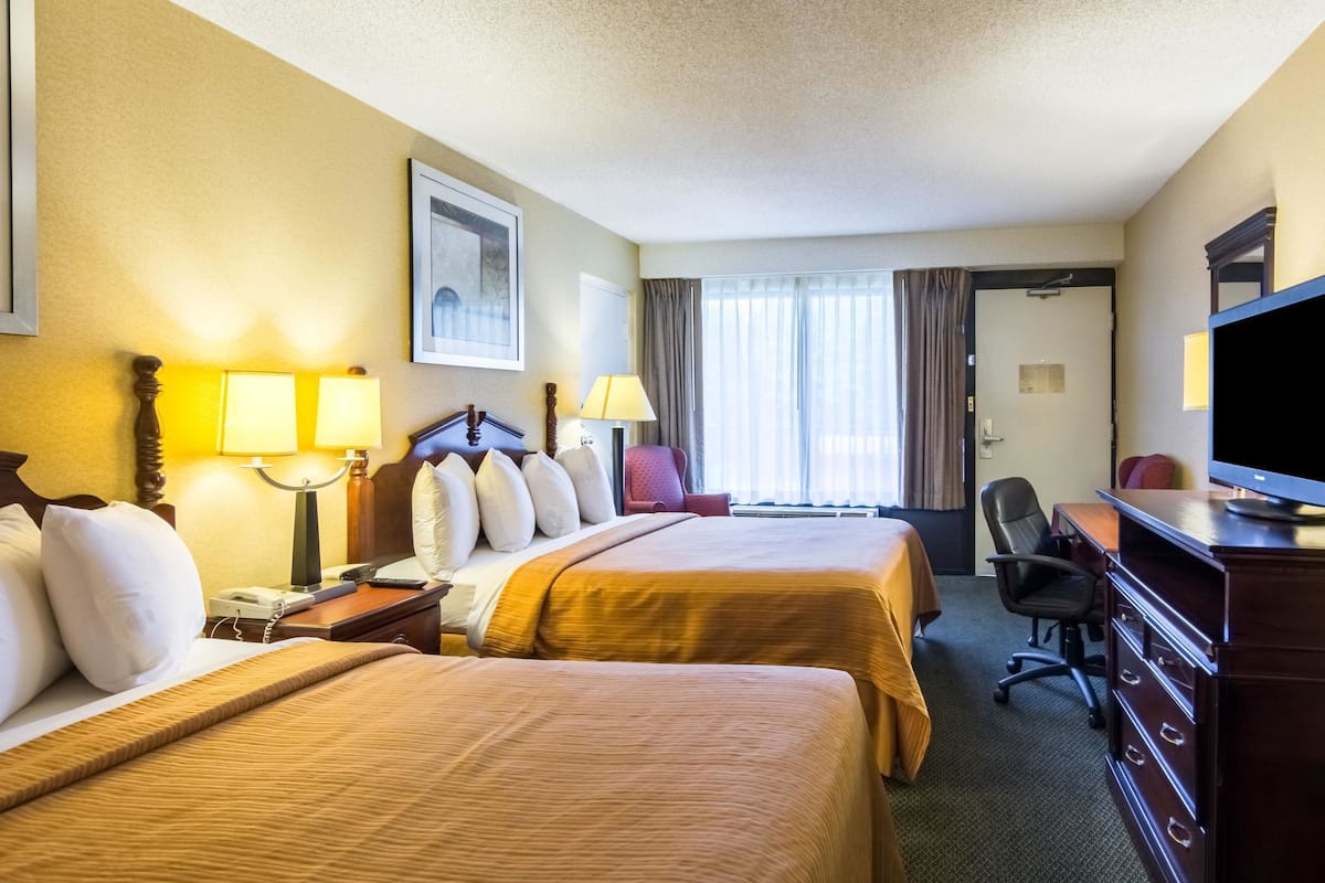 Quarto de hotel com duas camas, uma escrivaninha, uma cadeira, uma televisão e uma janela com cortinas.