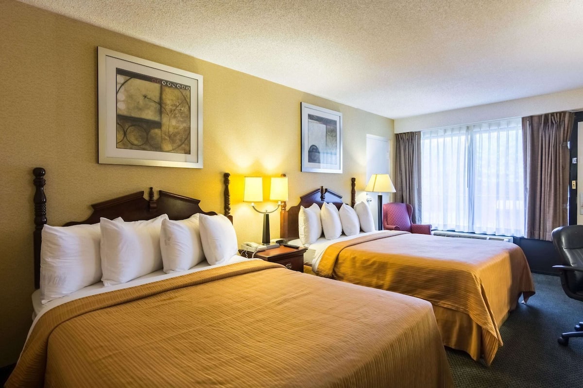Quarto de hotel com duas camas, uma escrivaninha, uma cadeira e uma janela com cortinas.