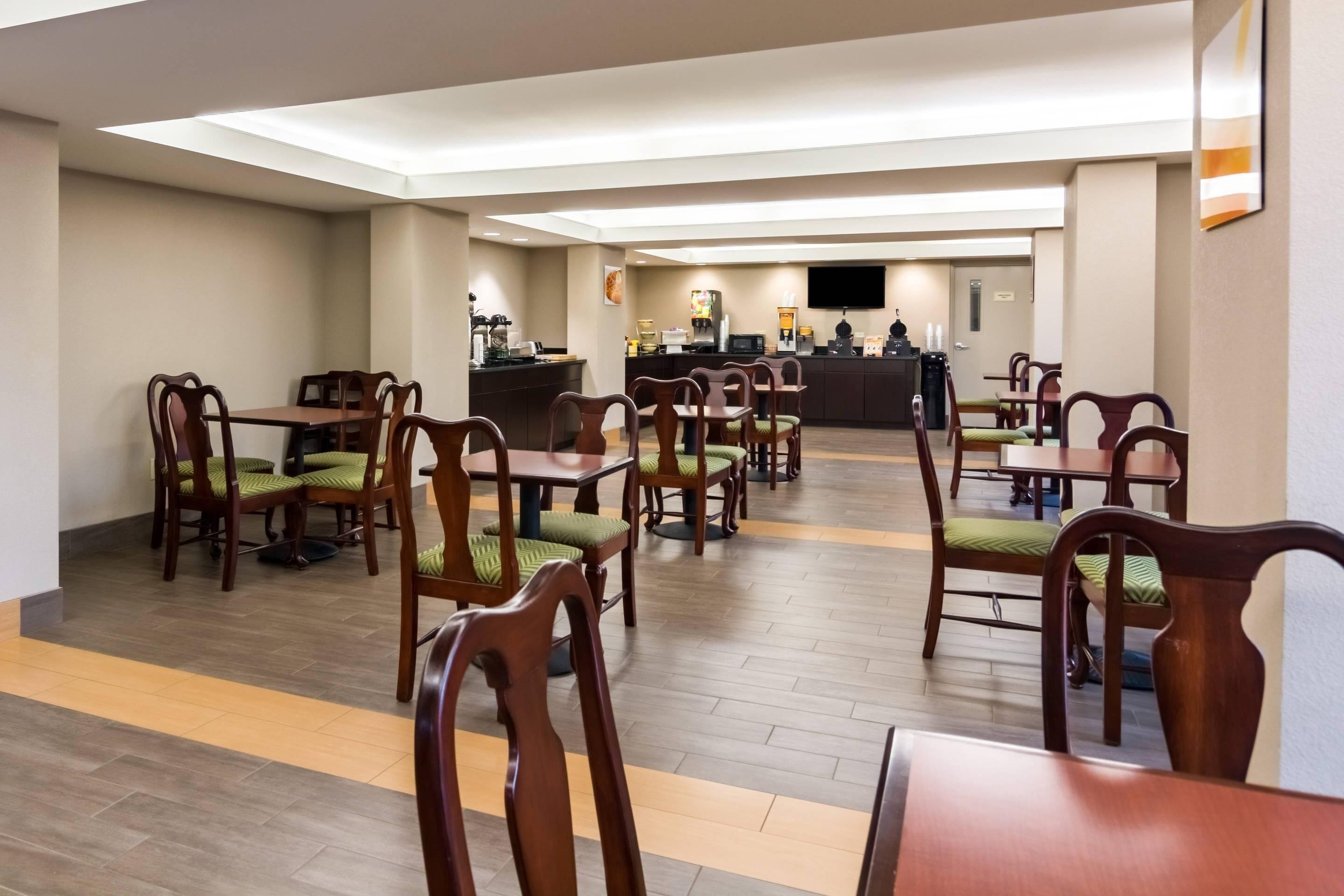 A hotel dining area with multiple tables and chairs, a central counter with a coffee machine, and a flat-screen TV mounted on the wall.