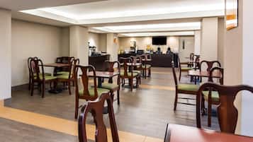 A hotel dining area with multiple tables and chairs, a central counter with a coffee machine, and a flat-screen TV mounted on the wall.