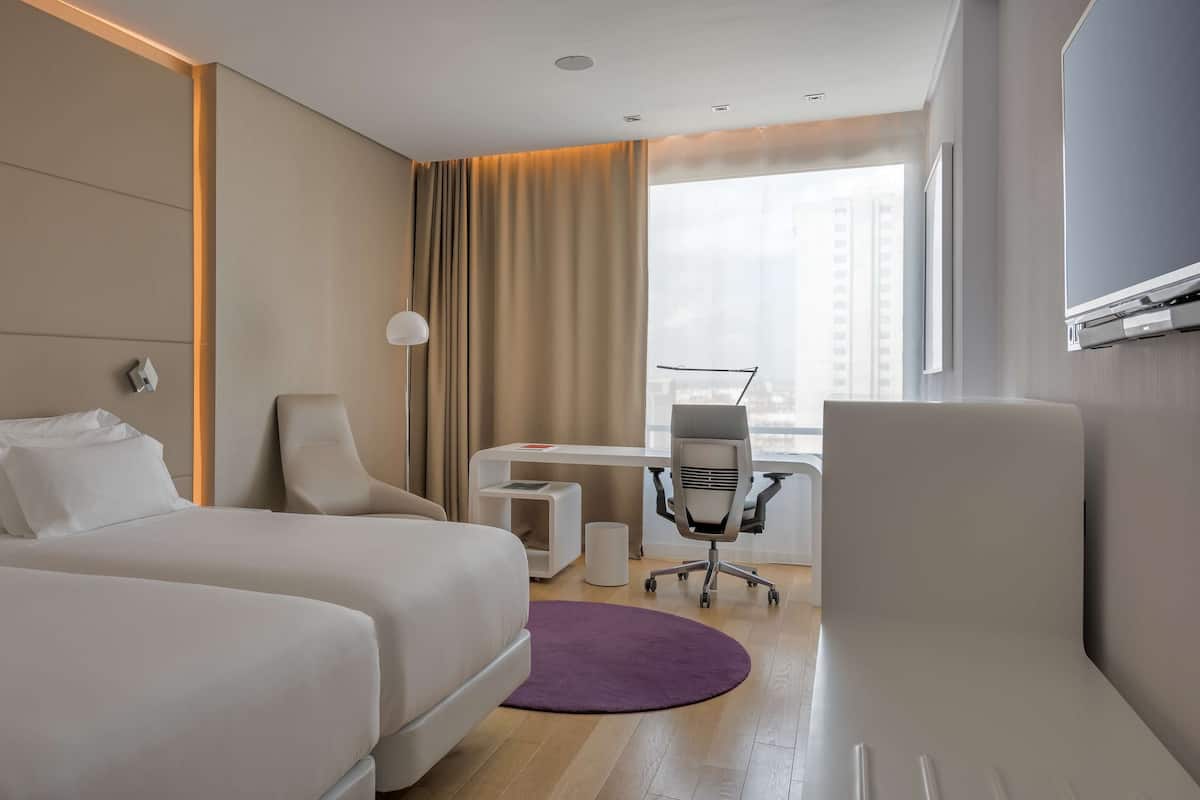 A hotel room with two beds, a desk, a chair, and a large window.