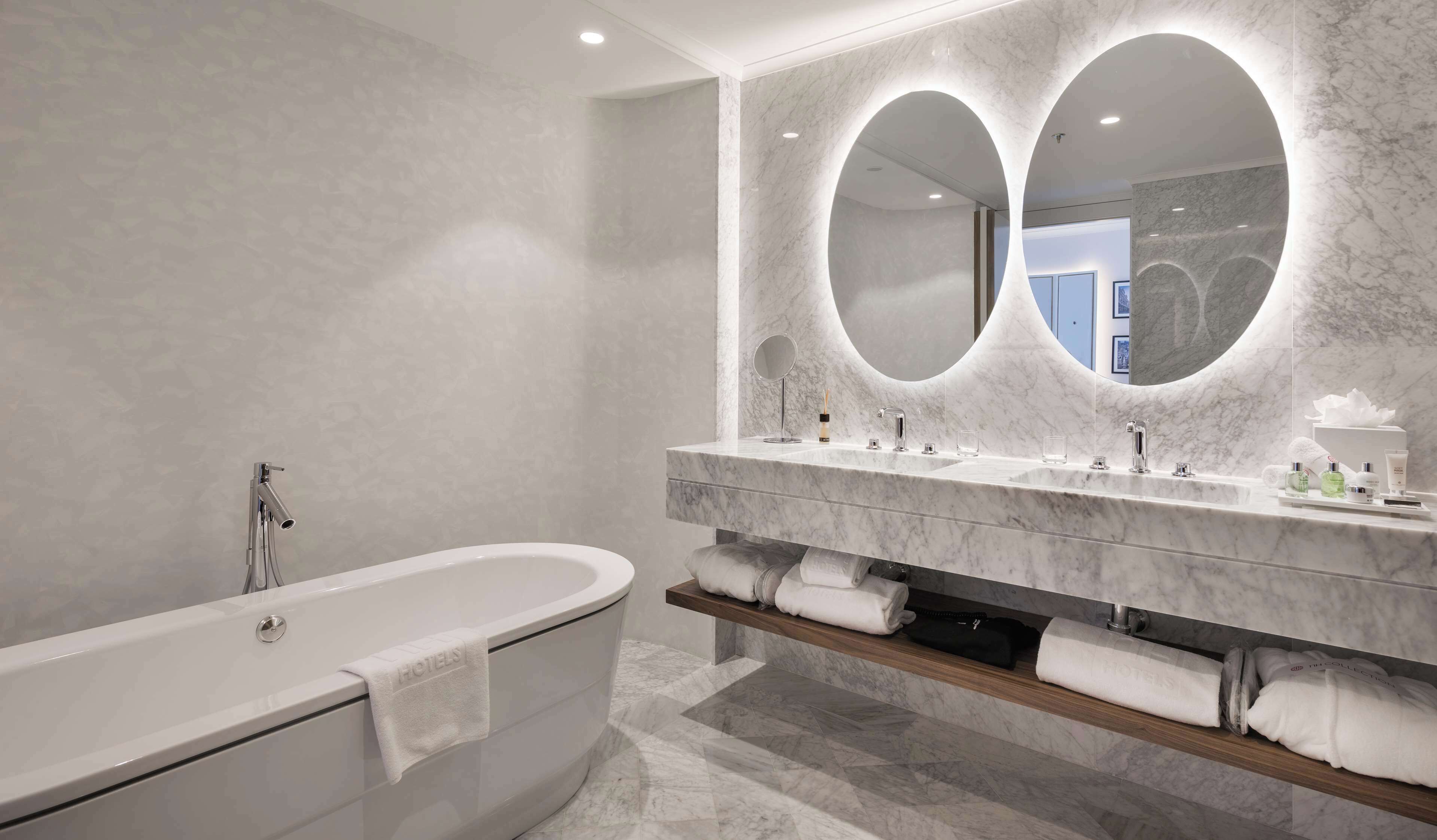 Un baño moderno con una bañera independiente, un espejo grande con dos luces ovaladas y un mueble de tocador con varios lavamanos y artículos de aseo.