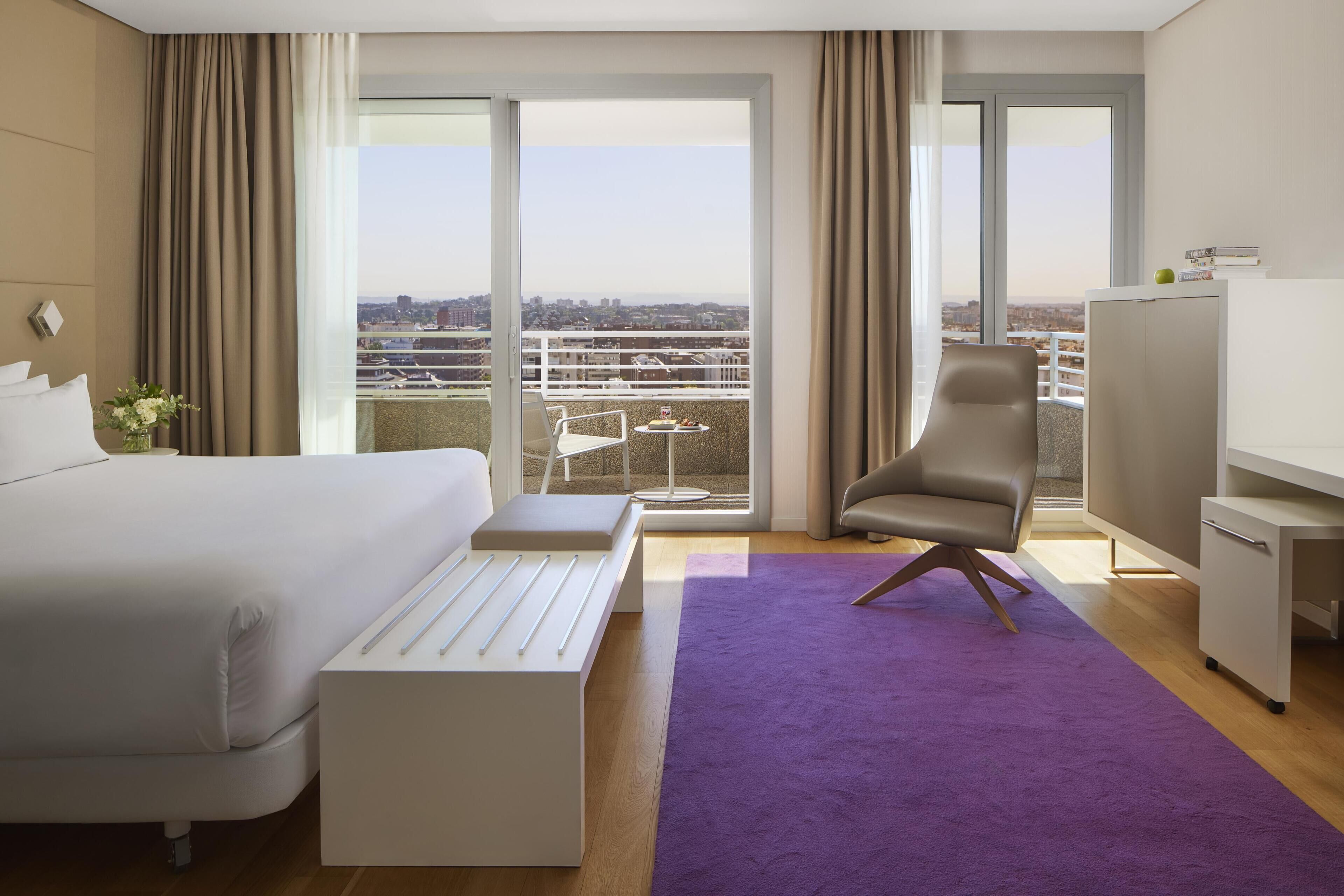 Ein Hotelzimmer mit Bett, Schreibtisch, Stuhl, einem violetten Teppich und Blick auf die Stadt durch große Fenster.