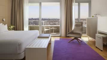 Ein Hotelzimmer mit Bett, Schreibtisch, Stuhl, einem violetten Teppich und Blick auf die Stadt durch große Fenster.