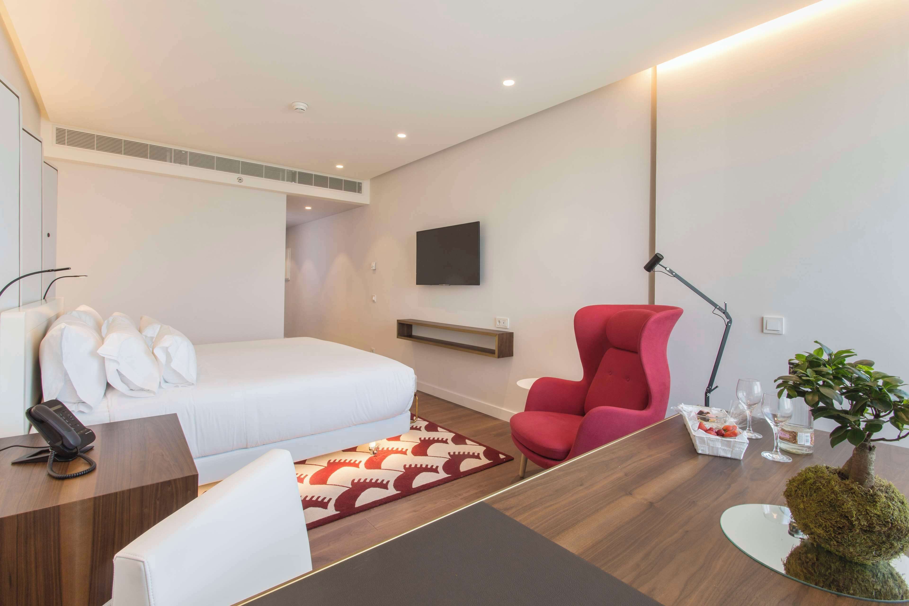 A modern hotel room with a bed, a desk with a telephone, a red chair, a small table with a plant, and a flat-screen TV mounted on the wall.