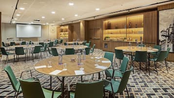Ein Konferenzraum mit mehreren Tischen und Stühlen, einer Projektionsleinwand sowie einer Barbereich mit Flaschen- und Glasregalen.