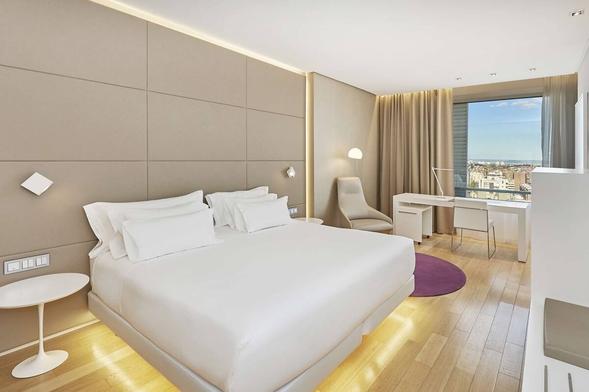 A modern hotel room with a large bed, a desk, a chair, a TV, and a city view.