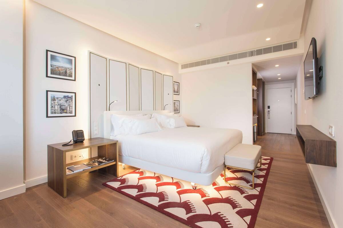 A modern hotel room with a large bed, a bedside table with a phone, a small desk, and a TV mounted on the wall.