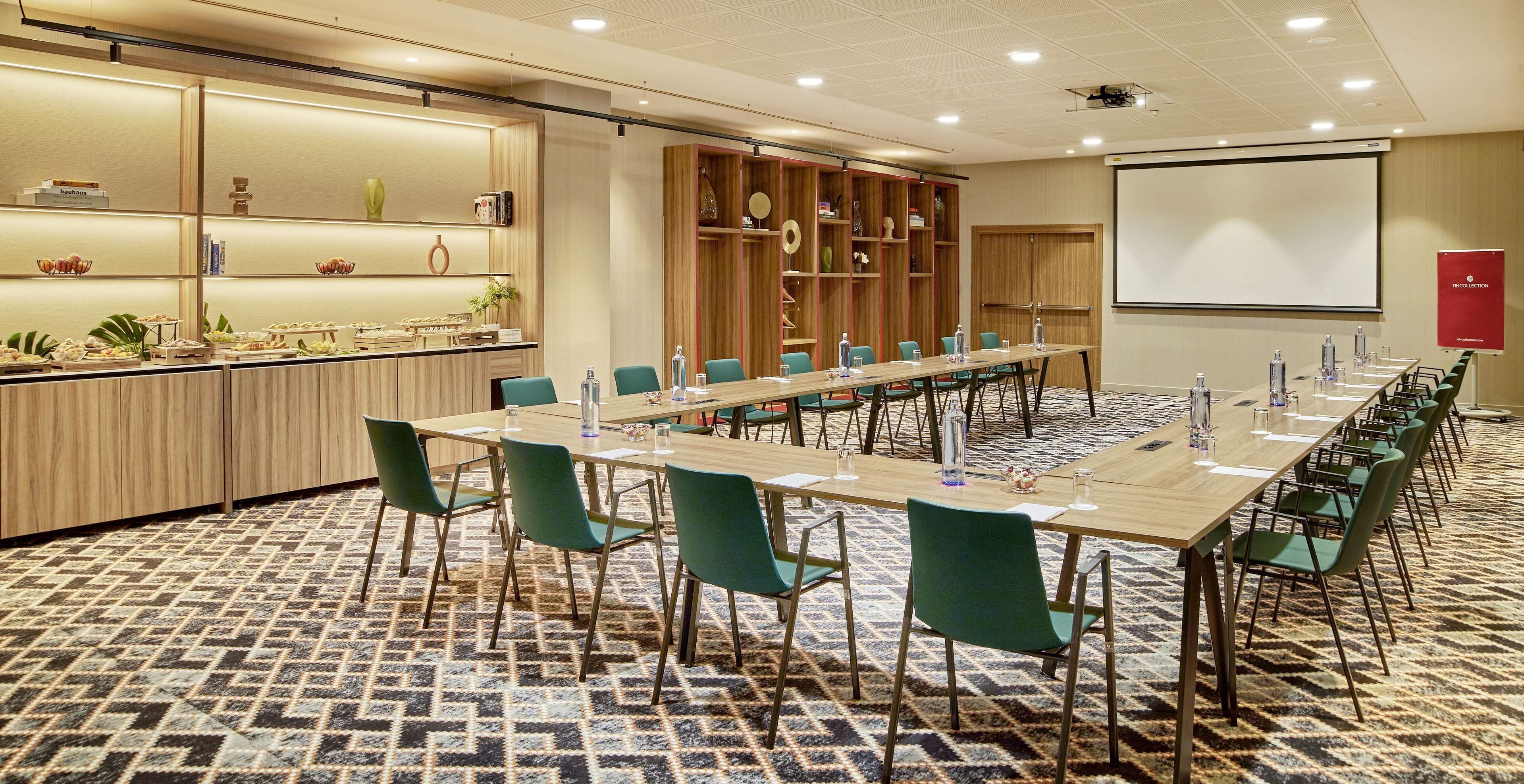 A conference room with a long table, green chairs, a projector screen, and a buffet area with various items.