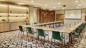 A conference room with a long table, green chairs, a projector screen, and a buffet area with various items.
