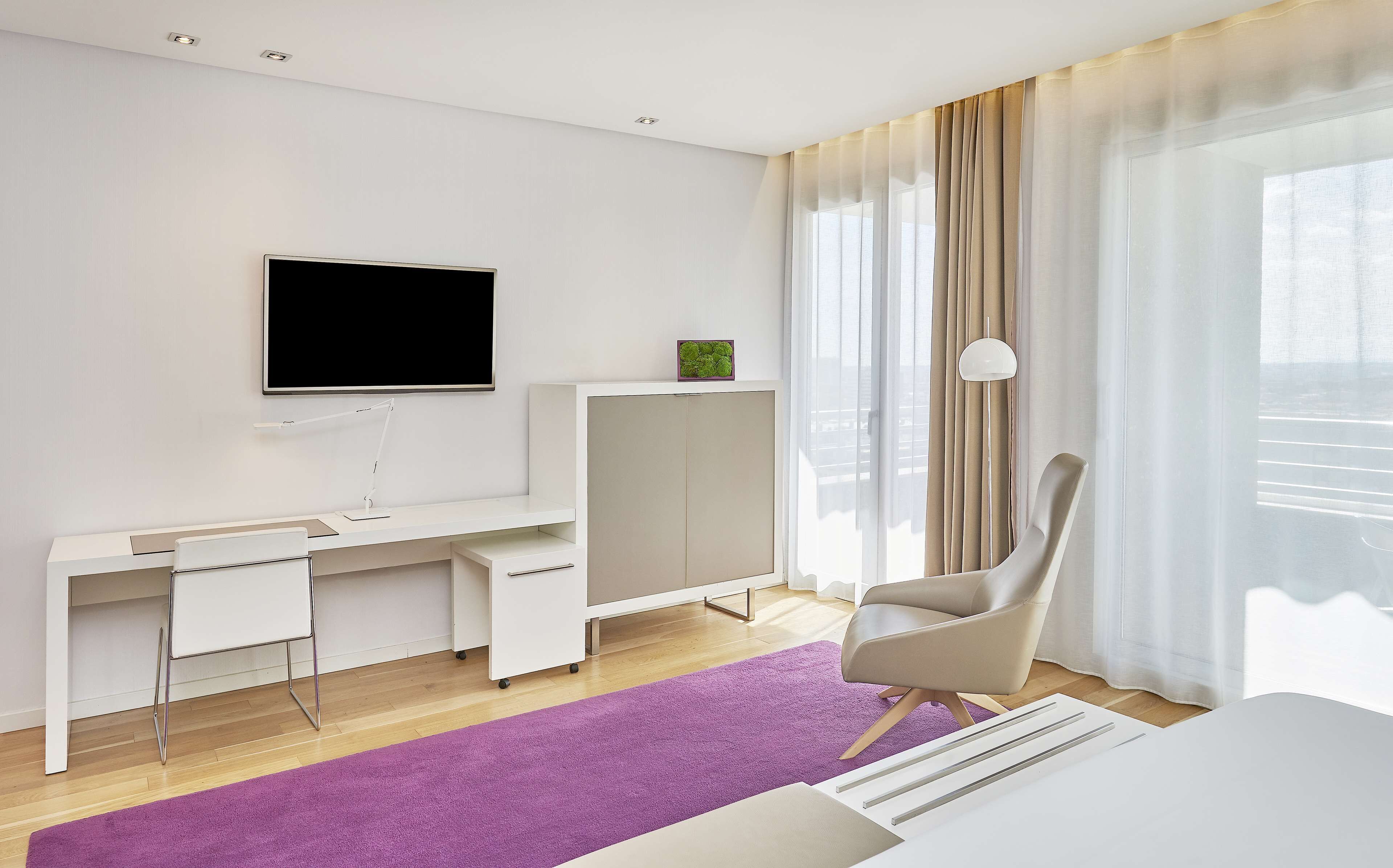 Quarto de hotel moderno com uma escrivaninha branca, televisão de tela plana, poltrona confortável e uma janela grande com cortinas.