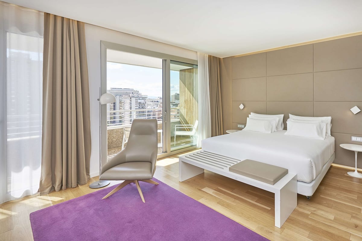 A modern hotel room with a large bed, a chair, a small table, and a view of the city through large windows.