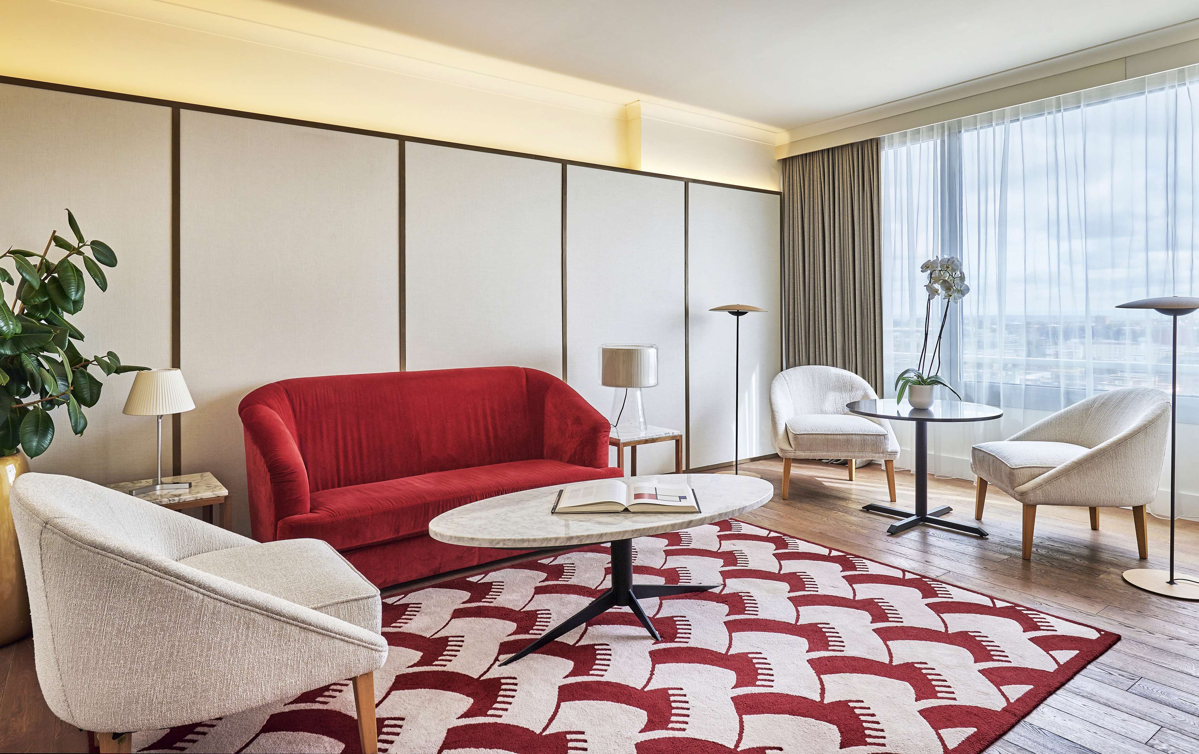 Une chambre d’hôtel moderne avec un canapé rouge, deux fauteuils blancs, une table basse ronde et un tapis à motifs.