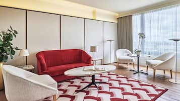 Une chambre d’hôtel moderne avec un canapé rouge, deux fauteuils blancs, une table basse ronde et un tapis à motifs.