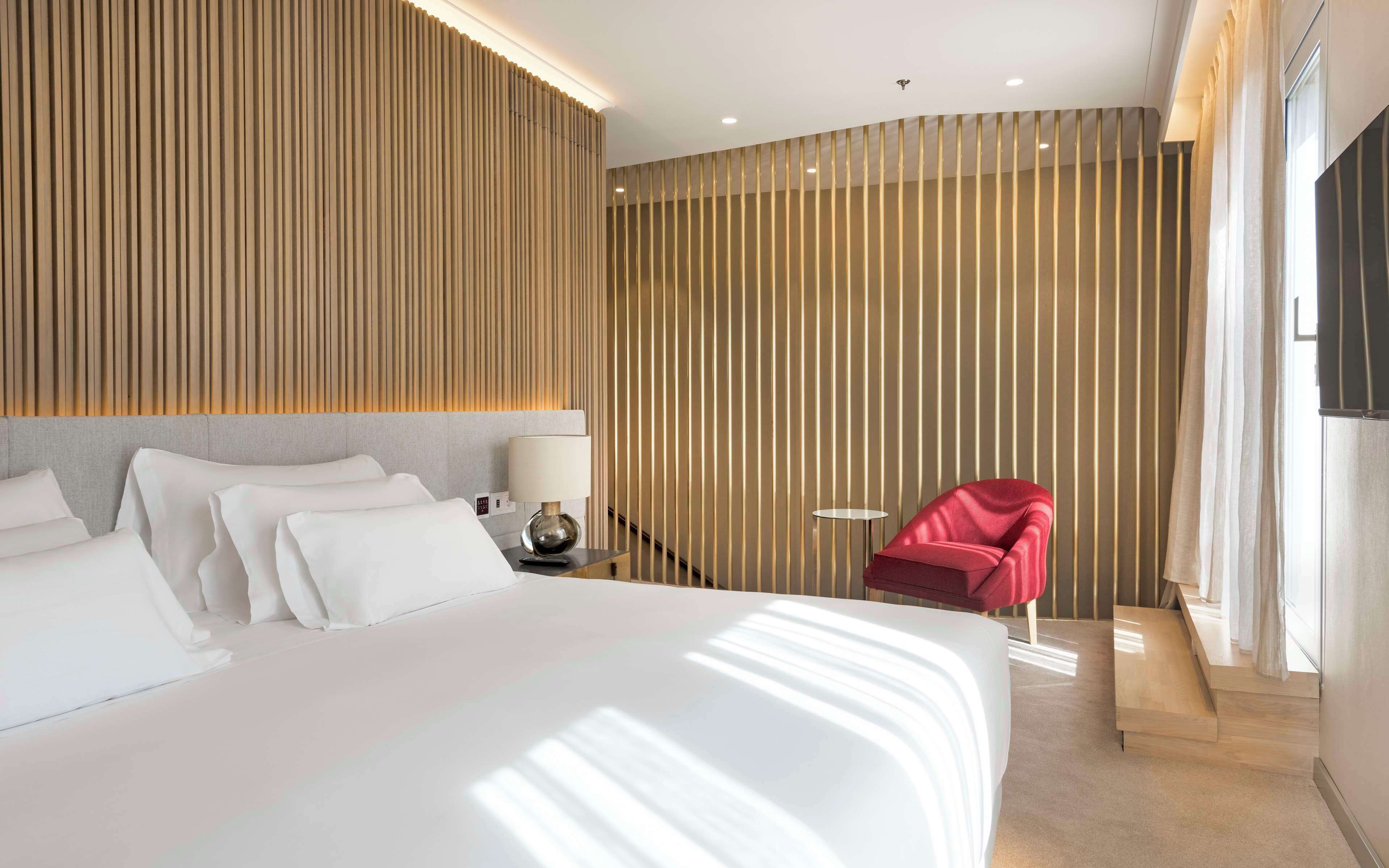 A modern hotel room with a large bed, a red armchair, and a view of the outdoors.