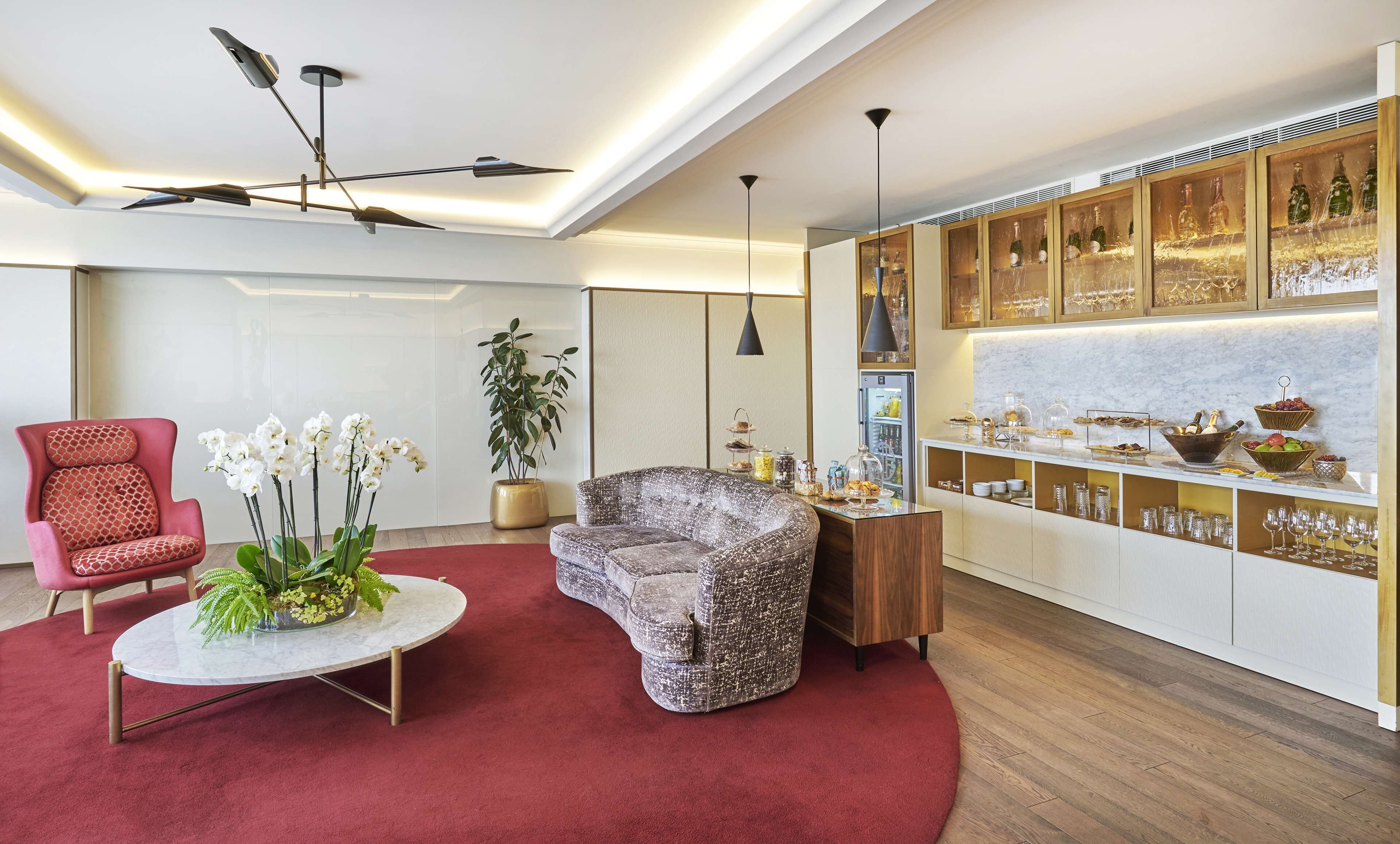 Una sala de estar moderna con alfombra roja, un sillón rojo, un sillón estampado, una mesa de centro redonda con una planta y un ventanal grande.