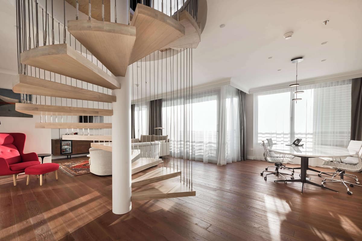 A spacious living area with a modern staircase, a red chair, a white sofa, a dining table with chairs, and large windows with sheer curtains.