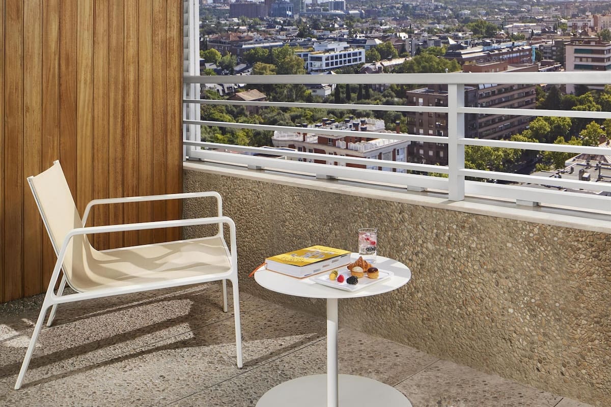 A balcony with a white chair, a small round table with snacks, and a city view.