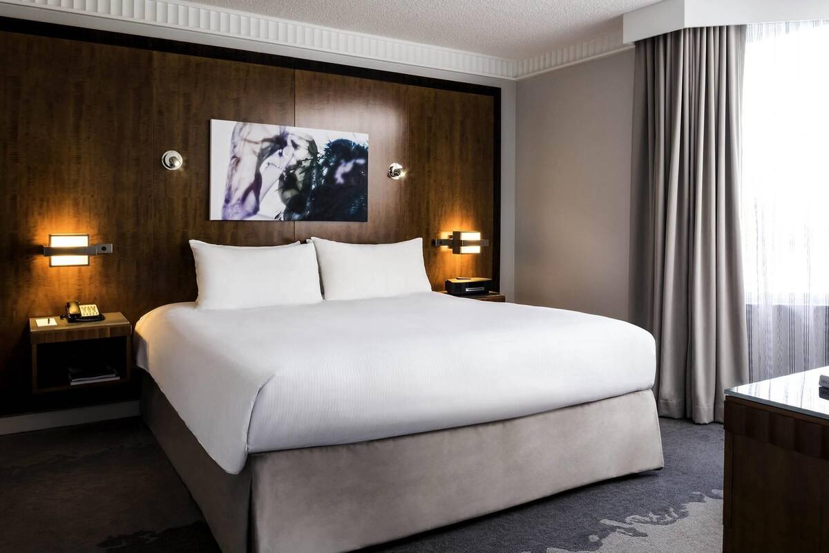 Hotellihuone, jossa on suuri sänky, kaksi yöpöytää valaisimineen, seinälle asennettu televisio ja ikkuna, jossa on verhot.