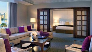 A modern hotel room with a sofa, armchair, and a coffee table. There is a bed with a nightstand and a lamp. The room has a large window with a view of the city. - Pullman Miami Airport (Miami)