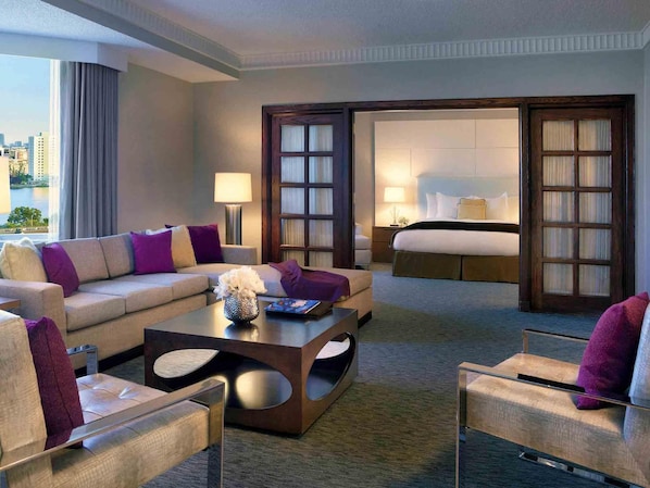 A modern hotel room with a sofa, armchair, and a coffee table. There is a bed with a nightstand and a lamp. The room has a large window with a view of the city.