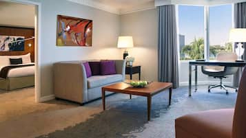 Habitación de hotel con cama, sofá, mesa de centro con fruta, escritorio y silla, lámpara y un cuadro en la pared.