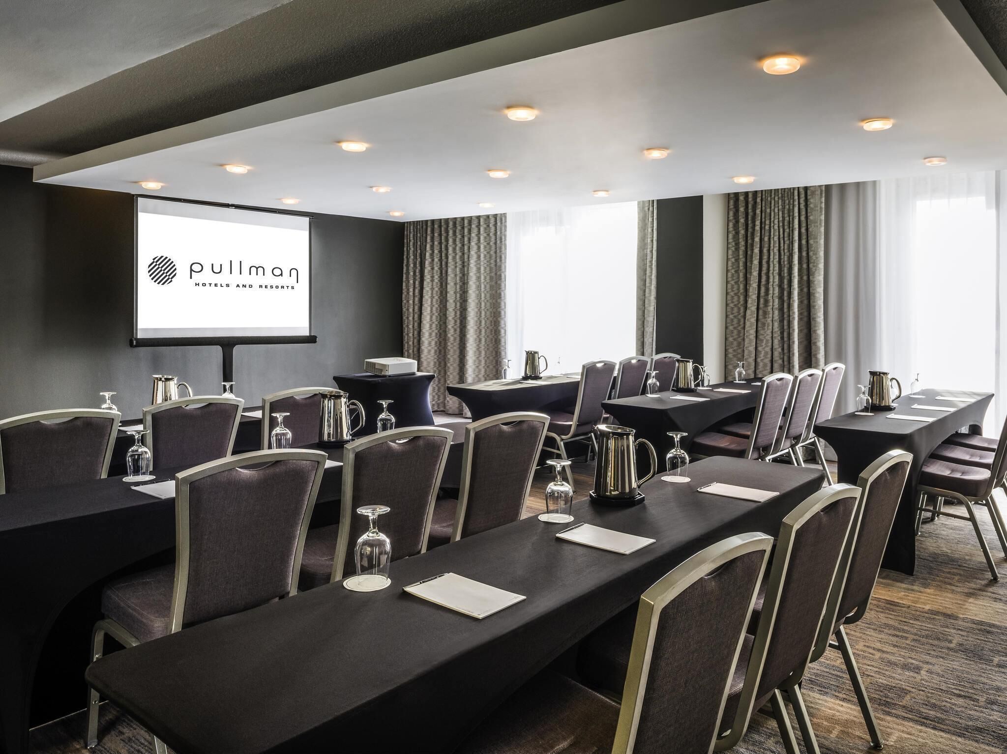 Un salón de conferencias con una pantalla de proyector que muestra el logotipo de 'Pullman Hotels and Resorts', varias mesas con sillas y suelo alfombrado.